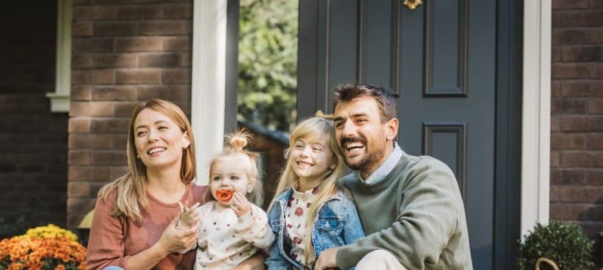 A family of four sitting on the front porch of their home in autumn light — mom and dad in casual clothes with two young kids, pumpkins and mums on the steps, an everyday lived-in moment.