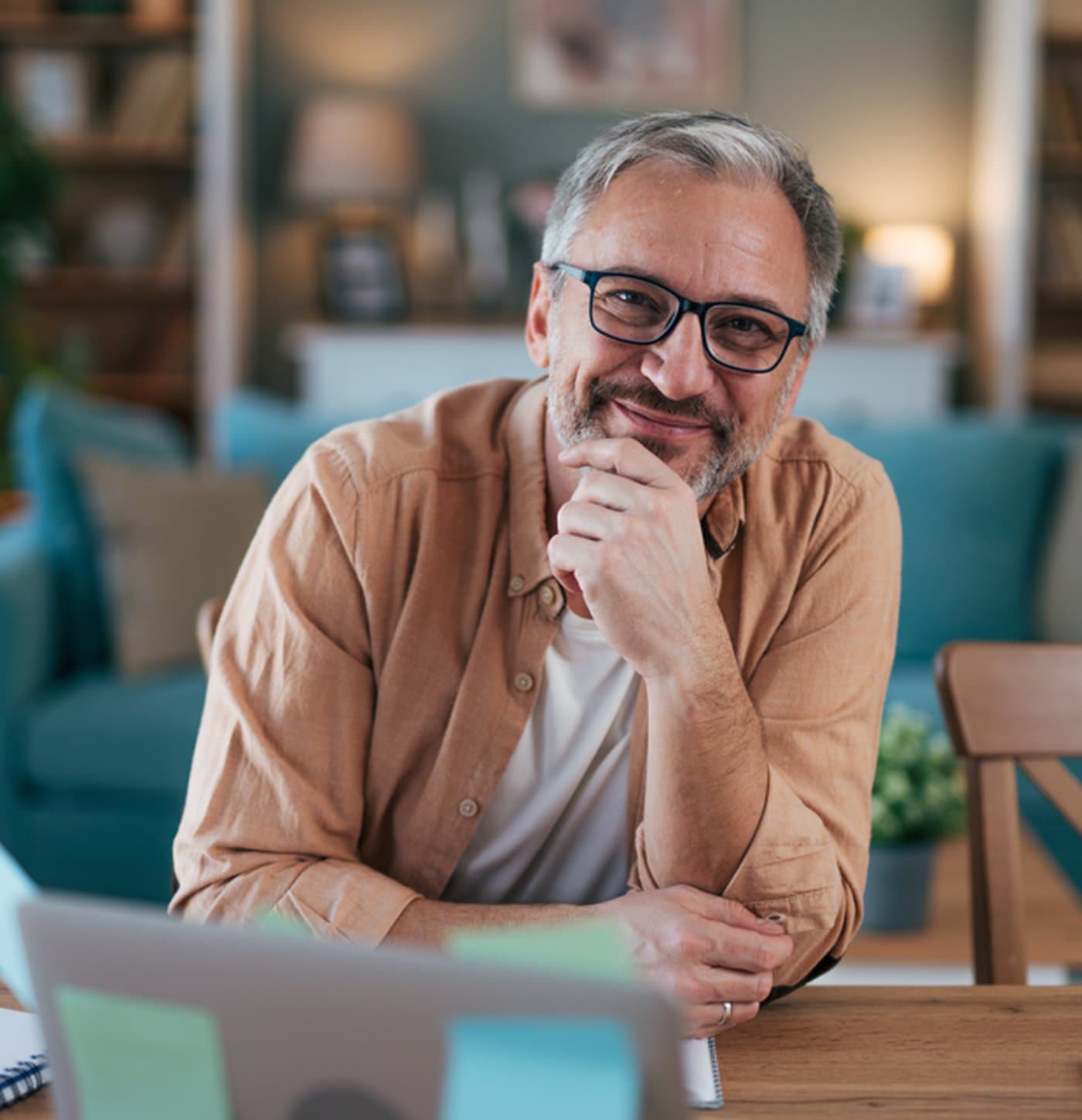 A person sitting at a home desk in front of a laptop in a softly lit living room — the kind of everyday moment when a money question shows up.