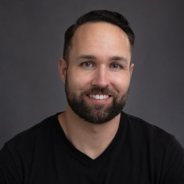 Allen Mueller, a smiling man with short dark hair and a beard, wearing a black v-neck shirt, photographed against a soft grey backdrop.